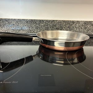 May include: A small, stainless steel frying pan with a copper-colored base and a black handle. The pan is sitting on a black electric stovetop. The handle has a loop for hanging.