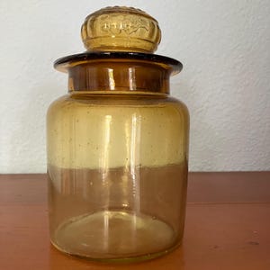May include: A clear amber glass jar with a textured lid. The jar has a round body and a wide mouth.