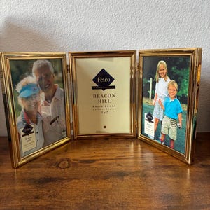 May include: A gold-colored, three-panel photo frame with pictures in two panels and a blank panel in the center. The frame is labeled "Fetco Beacon Hill Solid Brass" and is designed to hold 5x7 inch photos. The frame is on a wooden surface.