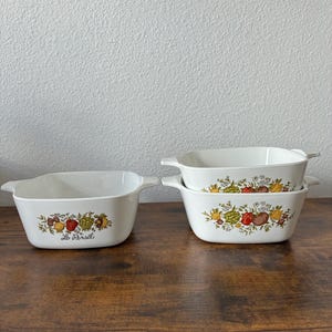 May include: White rectangular baking dishes with handles. Each dish features a colorful fruit and vegetable design. One dish has the text "Le Persil". The dishes are stacked on a wooden surface.