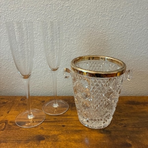 May include: Two clear champagne flutes and a crystal ice bucket with a gold rim and handles sit on a wooden surface. The ice bucket has a diamond pattern. The background is a plain, off-white wall.