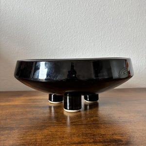 May include: A glossy black ceramic bowl with a wide, shallow design. The bowl is elevated by three short, cylindrical legs with white bases. The bowl sits on a dark brown wooden surface, reflecting the light.