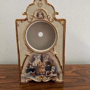 May include: A decorative ceramic clock with a cream-colored background and gold trim. The clock features a circular opening and a painted scene of several kittens. A small kitten figurine sits at the top.