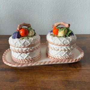 May include: A white ceramic salt and pepper set with a decorative fruit design. The set includes two lidded jars with a woven pattern and a matching tray. Each lid features a cluster of grapes, a tomato, and other fruits.