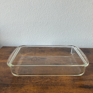 May include: A clear, rectangular glass baking dish with rounded handles on each end. The dish is empty and sits on a dark wooden surface. The background is a plain white wall.