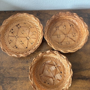 May include: Three round, shallow woven baskets made of light brown natural reeds. Each basket features a complex, geometric lattice pattern at the base and a thick, braided rim. The baskets are arranged in a triangular formation on a flat surface.
