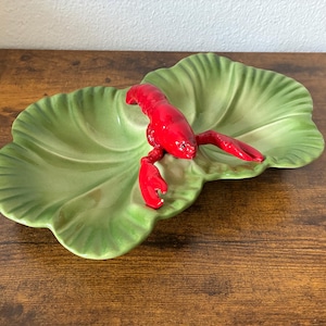 May include: A ceramic serving dish shaped like a large green lettuce leaf with a bright red lobster figure attached to the center. The dish features a glossy finish and realistic molded details on both the leaf and the crustacean.