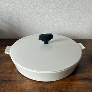 May include: A white, round casserole dish with a lid and a black handle. The dish has two small handles on either side. The lid is centered with a black handle. The dish is sitting on a wooden surface.