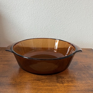May include: A round, amber-colored glass baking dish with two handles. The dish has a smooth, curved design and is sitting on a wooden surface. The dish is empty and appears to be clean.