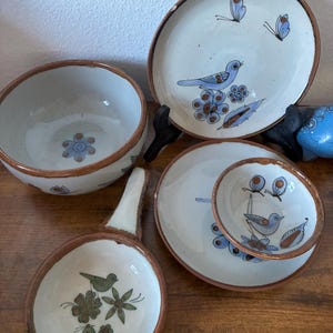 May include: A collection of ceramic tableware featuring off-white surfaces with brown rims. The pieces include a bowl, two plates, and a small handled dish, all decorated with hand-painted blue and green birds, butterflies, and floral motifs.