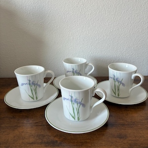 May include: A set of four white teacups and saucers. Each cup and saucer features a delicate floral design with purple irises and green stems. The saucers have a green trim, and the cups have a curved handle.