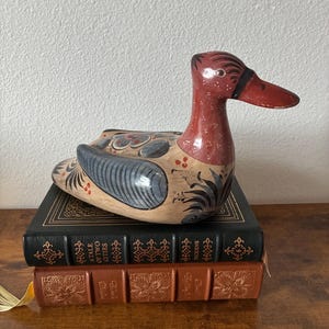 May include: A hand-painted wooden duck figurine featuring a reddish-brown head and a cream-colored body decorated with blue-grey wings, black floral accents, and small red dots, resting on two stacked hardcover books titled A Tale of Two Cities.