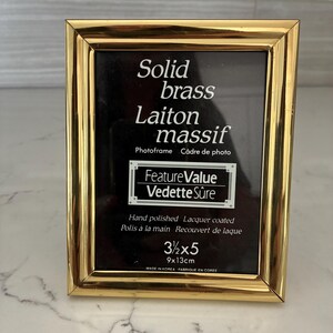 May include: A rectangular solid brass photo frame with a polished gold finish. The black insert features white text reading Solid brass, Laiton massif, Photoframe, Câdre de photo, FeatureValue, Vedette Sûre, Hand polished, Lacquer coated, 3 1/2 x 5, 9x13cm.