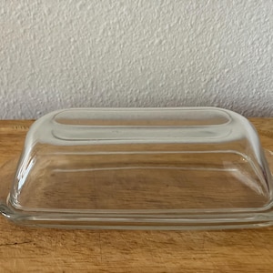 May include: A clear glass butter dish with a rectangular base and a matching lid. The dish is sitting on a wooden cutting board, and the background is a white wall.