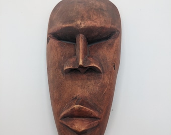 Pode incluir: Uma máscara de madeira esculpida castanha com uma expressão serena. A máscara apresenta olhos fechados, um nariz proeminente e lábios cheios. A superfície tem uma aparência texturizada, sugerindo um item feito à mão. A máscara é provavelmente para fins decorativos.