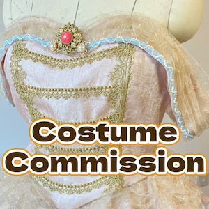 May include: A pink and gold bodice with a blue trim and a large gold brooch with a red stone. The bodice is part of a costume and the text "Costume Commission" is superimposed on the image.