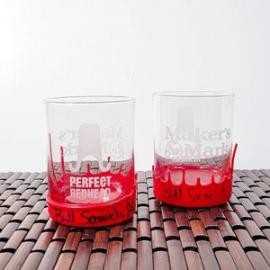 May include: Two clear glass tumblers with red liquid dripping down the sides. The tumbler on the left has the text "Perfect Redhead" written on the bottom. The tumbler on the right has the text "Maker's Mark" written on the bottom. Both tumblers are sitting on a brown bamboo mat.