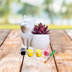 May include: White ceramic cat planter with a purple succulent plant inside. The planter is sitting on a wooden surface with four small paint pots and a paintbrush.