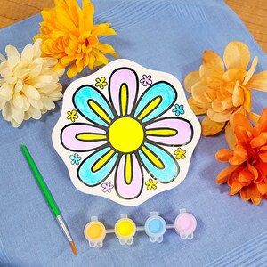 May include: A white ceramic plate with a colorful flower design. The flower has pink, blue, and yellow petals and a yellow center. The plate is surrounded by orange and yellow flowers.
