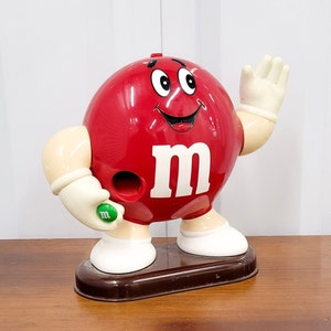 May include: Red M&M candy character figurine with a white smile, white eyes, and a white "M" on its chest. The figurine is holding a green M&M in its right hand and has a white glove on its left hand. The figurine is standing on a brown base.
