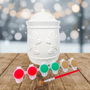 May include: A white ceramic cookie jar with a lid, featuring embossed gingerbread men. Includes red, green, and brown paint pots, and a small paintbrush with a red handle. The jar is on a wooden surface.