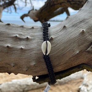 Op de afbeelding: Een zwarte gevlochten armband met een witte schelpbedel. De armband ligt op een tak.