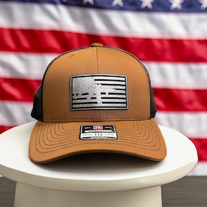 Könnte beinhalten: Eine braune und schwarze Baseballkappe mit einem schwarz-weißen amerikanischen Flaggen-Patch und einem roten, weißen und schwarzen Richardson 112 verstellbaren Etikett.