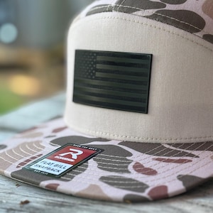 Op de afbeelding: Een flat bill snapback pet met een camouflagepatroon in bruin, beige en roze tinten. De pet heeft een zwart Amerikaans vlagpatch en een "Richardson" label. Gemaakt van duurzame stof.