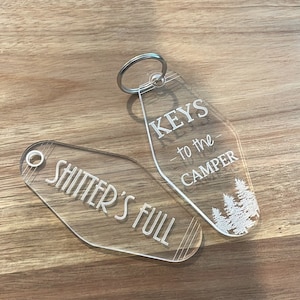 May include: Two clear acrylic keychains with white text. One keychain says "Keys to the Camper" with a small image of pine trees. The other keychain says "Shitter's Full".