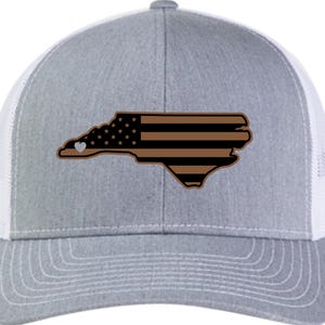 May include: A grey and white trucker hat with a brown and black American flag design in the shape of the state of North Carolina. A small white heart is in the top left corner of the state outline.