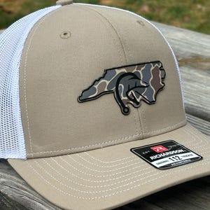 Może przedstawiać: Beżowo-biała czapka typu trucker z naszywką w kamuflażu w kształcie Karoliny Północnej. Naszywka przedstawia wzór haczyka wędkarskiego. Czapka ma zakrzywiony daszek i siatkowy tył. Etykieta marki głosi "Richardson".