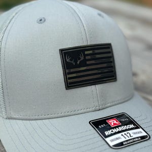 Könnte beinhalten: Eine graue und schwarze Trucker-Kappe mit einem Tarnmuster-Patch der amerikanischen Flagge mit einem weißen Hirschgeweih-Design. Die Kappe hat einen schwarzen verstellbaren Riemen mit dem Text "Richardson 112 Adjustable" und "EST. 1970".