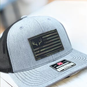 Può includere: Un cappello da camionista grigio e nero con una toppa a forma di bandiera americana mimetica che presenta una silhouette di cervo. Il cappello ha una parte posteriore in rete nera e un'etichetta del marchio Richardson.