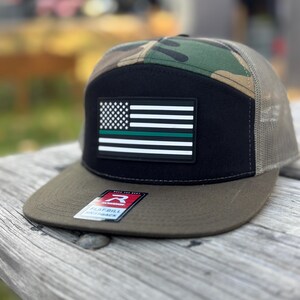 May include: A flat bill snapback hat with a camouflage top and a black front panel. The front panel features an American flag patch with a green stripe. The brim is olive green, and the mesh back is tan.