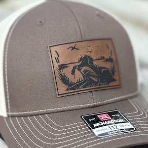 May include: A brown and white trucker cap with a leather patch depicting a dog in a field with ducks. The cap has a curved peak, white stitching, and an adjustable snap closure. The patch features a detailed illustration of a hunting dog. The cap is a Richardson 112 Trucker.