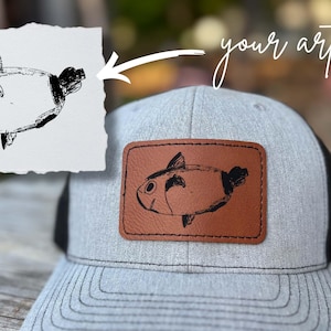 Puede incluir: Gorra de camionero gris con la parte trasera de malla negra y un parche de cuero marrón. El parche presenta una ilustración de pez en blanco y negro. Las palabras "your artwork" están escritas en cursiva arriba.