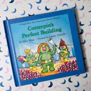May include: A children's book titled "Cotterpin's Perfect Building" featuring illustrations of green, cartoon-like characters. The book has a blue cover with the title in large, green letters. The book is a Fraggle Rock book starring Jim Henson's Muppets.