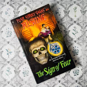 May include: A paperback book titled "The Sign of Fear" by R.L. Stine. The cover features a skull with glowing green eyes, a couple, and a pendant. The book is part of the "Fear Street Sagas" series.