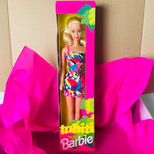 May include: A vintage Barbie doll in a pink and yellow box. The doll is wearing a colourful geometric print dress and has blonde hair. The box says "Tahiti Barbie".