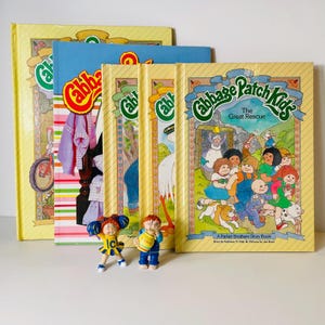 Puede incluir: Una colección de libros vintage de Cabbage Patch Kids, con cubiertas amarillas y el logotipo de la serie. Los libros están acompañados de dos pequeñas figuras de Cabbage Patch Kids. El título "The Great Rescue" es visible.