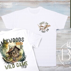 May include: Two white t-shirts with hunting-themed designs. The front of one shirt features a deer antler design with the text "Track 'em & Trap 'em". The back of the other shirt features a cabin in the woods with the text "Backwoods Bred & Wild Game Fed".