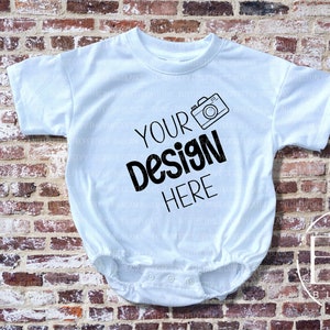 May include: A light blue baby onesie with a camera graphic and the text "YOUR DESIGN HERE". The onesie is lying on a brick wall background.