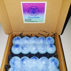 Op de afbeelding: Een bruine kartonnen doos met twee sets lichtblauwe wax melts. Elke set heeft een bubbelachtig ontwerp met donkerblauwe spikkels. De doos heeft een label met de tekst "Zuna Meltz" en social media.