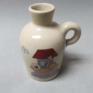 May include: A small, beige ceramic pitcher with a handle. The pitcher has a painted design of a red roofed house with a blue door and windows. There are animals on the roof of the house, including a giraffe, a sheep, and a lion.