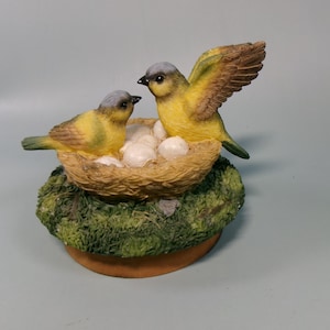 Peut inclure: Figurine d&eacute;corative repr&eacute;sentant deux oiseaux jaunes &agrave; t&ecirc;te grise et ailes marron. L'un d'eux a une aile d&eacute;ploy&eacute;e. Ils sont pos&eacute;s sur un nid avec des &oelig;ufs blancs, sur une base moussue et un socle circulaire brun.