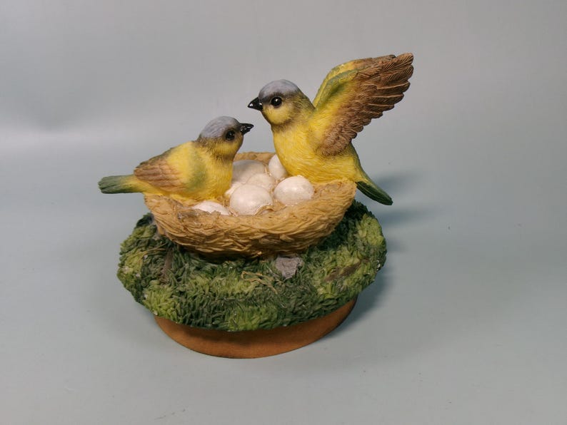 Peut inclure: Figurine d&eacute;corative repr&eacute;sentant deux oiseaux jaunes &agrave; t&ecirc;te grise et ailes marron. L'un d'eux a une aile d&eacute;ploy&eacute;e. Ils sont pos&eacute;s sur un nid avec des &oelig;ufs blancs, sur une base moussue et un socle circulaire brun.