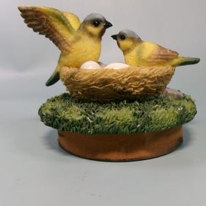 Peut inclure: Figurine d&eacute;corative repr&eacute;sentant deux oiseaux jaunes &agrave; t&ecirc;te grise, perch&eacute;s pr&egrave;s d'un nid avec des &oelig;ufs. Les oiseaux sont sur une base recouverte d'herbe verte artificielle et d'un bord marron.