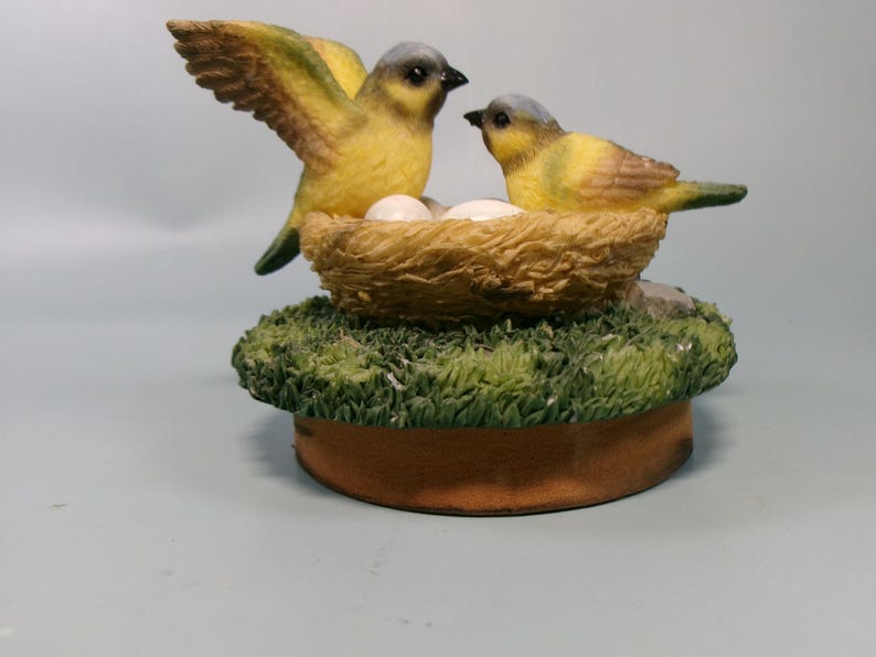Peut inclure: Figurine d&eacute;corative repr&eacute;sentant deux oiseaux jaunes &agrave; t&ecirc;te grise, perch&eacute;s pr&egrave;s d'un nid avec des &oelig;ufs. Les oiseaux sont sur une base recouverte d'herbe verte artificielle et d'un bord marron.