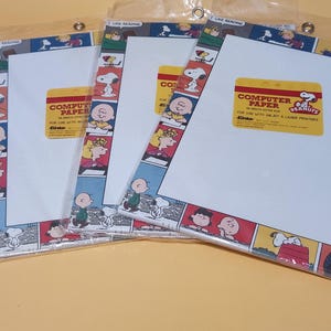 May include: Three packs of Peanuts computer paper in clear plastic sleeves. Each pack features a border of comic strip panels with Snoopy and friends. A yellow label on each pack reads Computer Paper, 50 Sheets Letter Size, Peanuts.