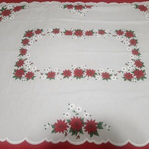 May include: A rectangular white tablecloth featuring a scalloped edge and a repeating pattern of red poinsettia flowers with green leaves and small white blossoms. The floral design is arranged in a central rectangular border and corners.